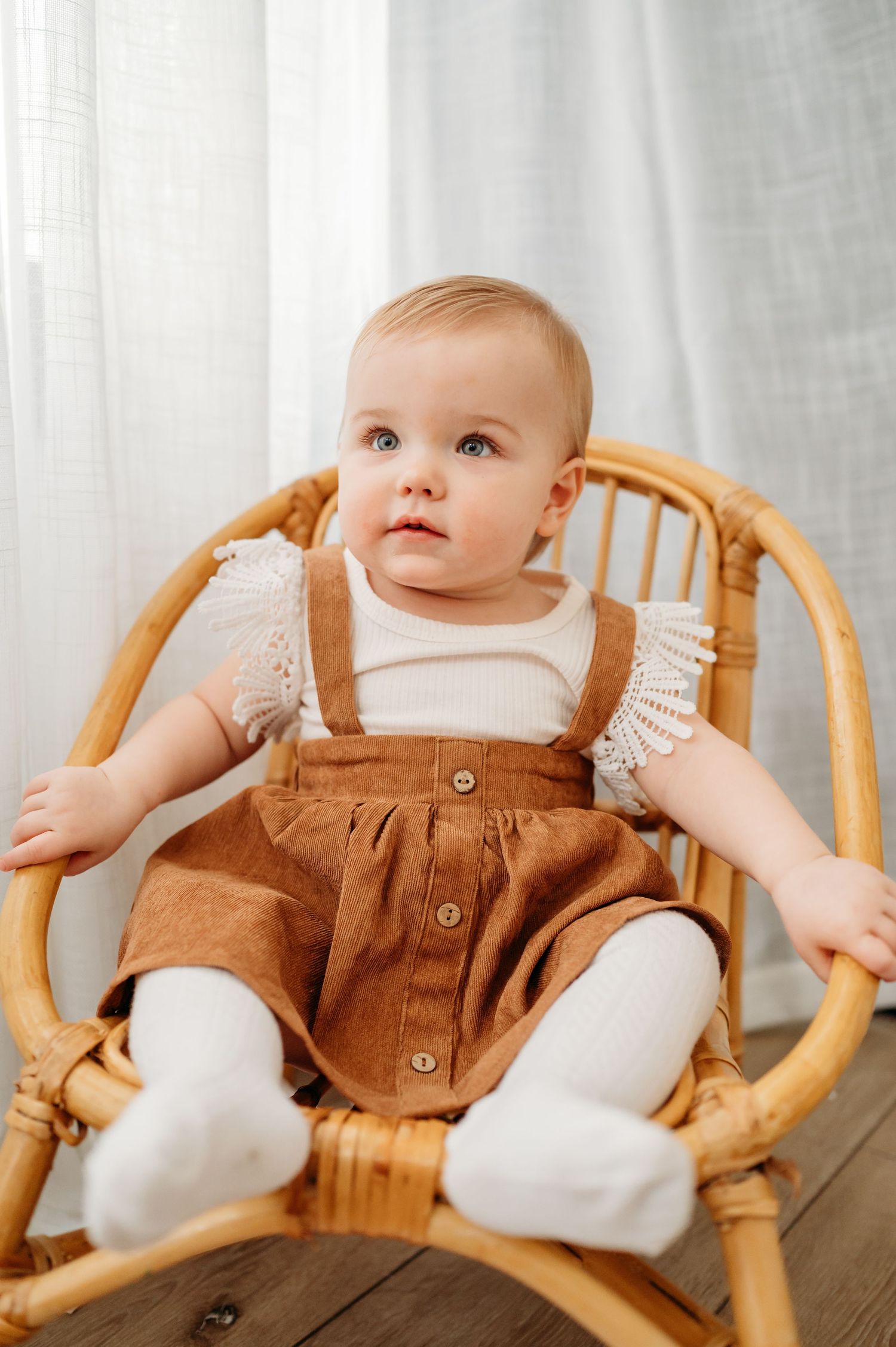 Sweet baby in brown corduroy overalls and white shirt sitting in a rattan chair against light backdrop.