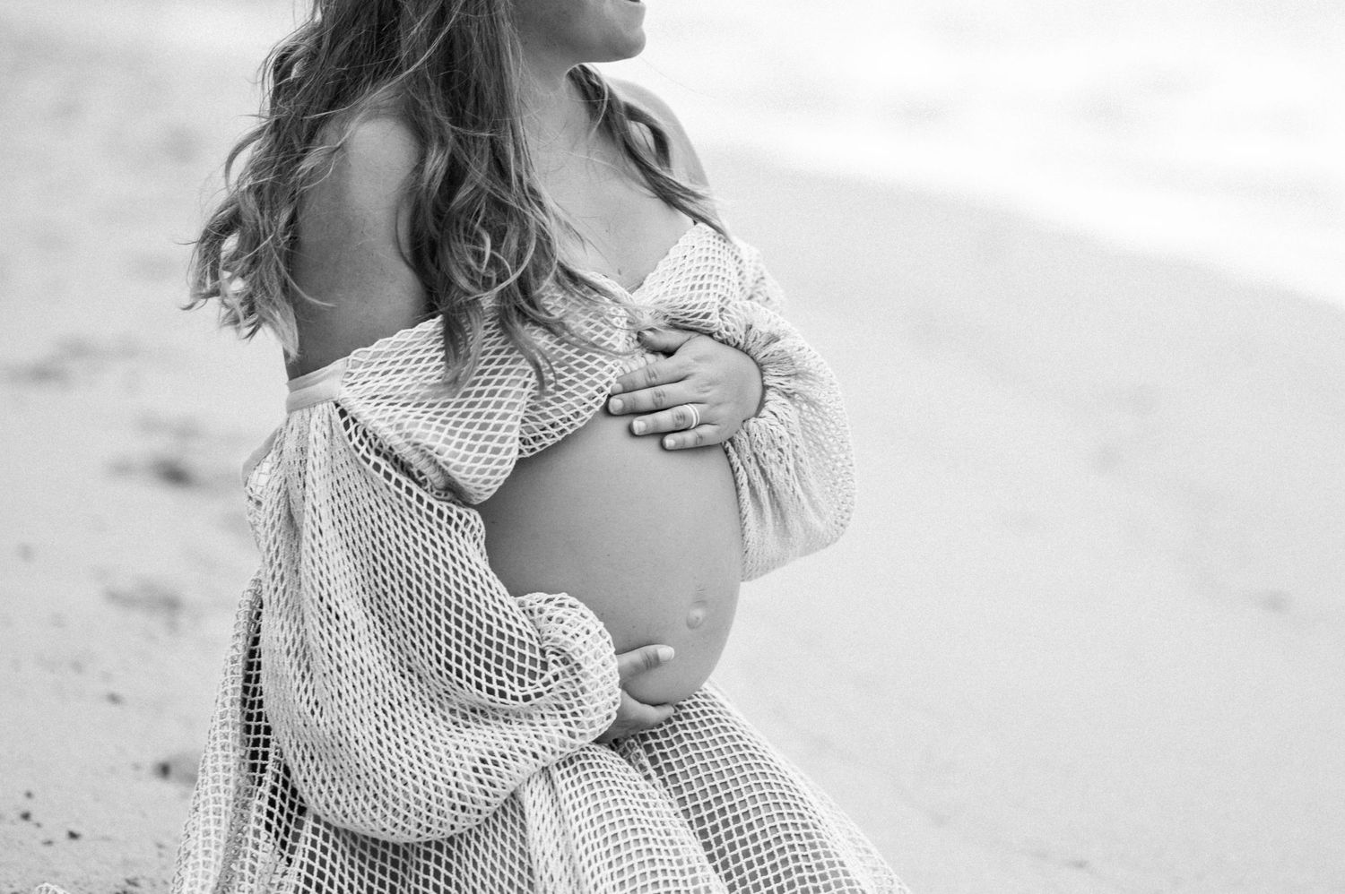 A pregnant woman in a flowy mesh cover up cradles her baby bump during a black and white beach maternity photoshoot.