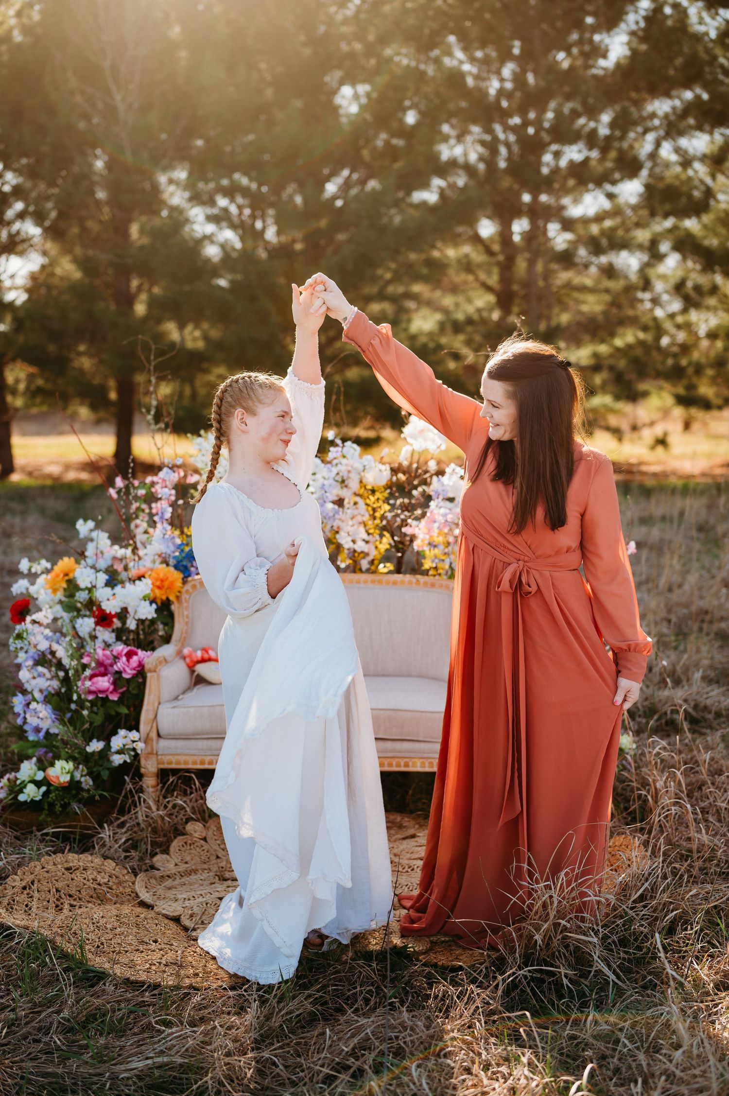 Two people in white and rust-colored dresses sit on a bench at sunset with colorful floral arrangements nearby.