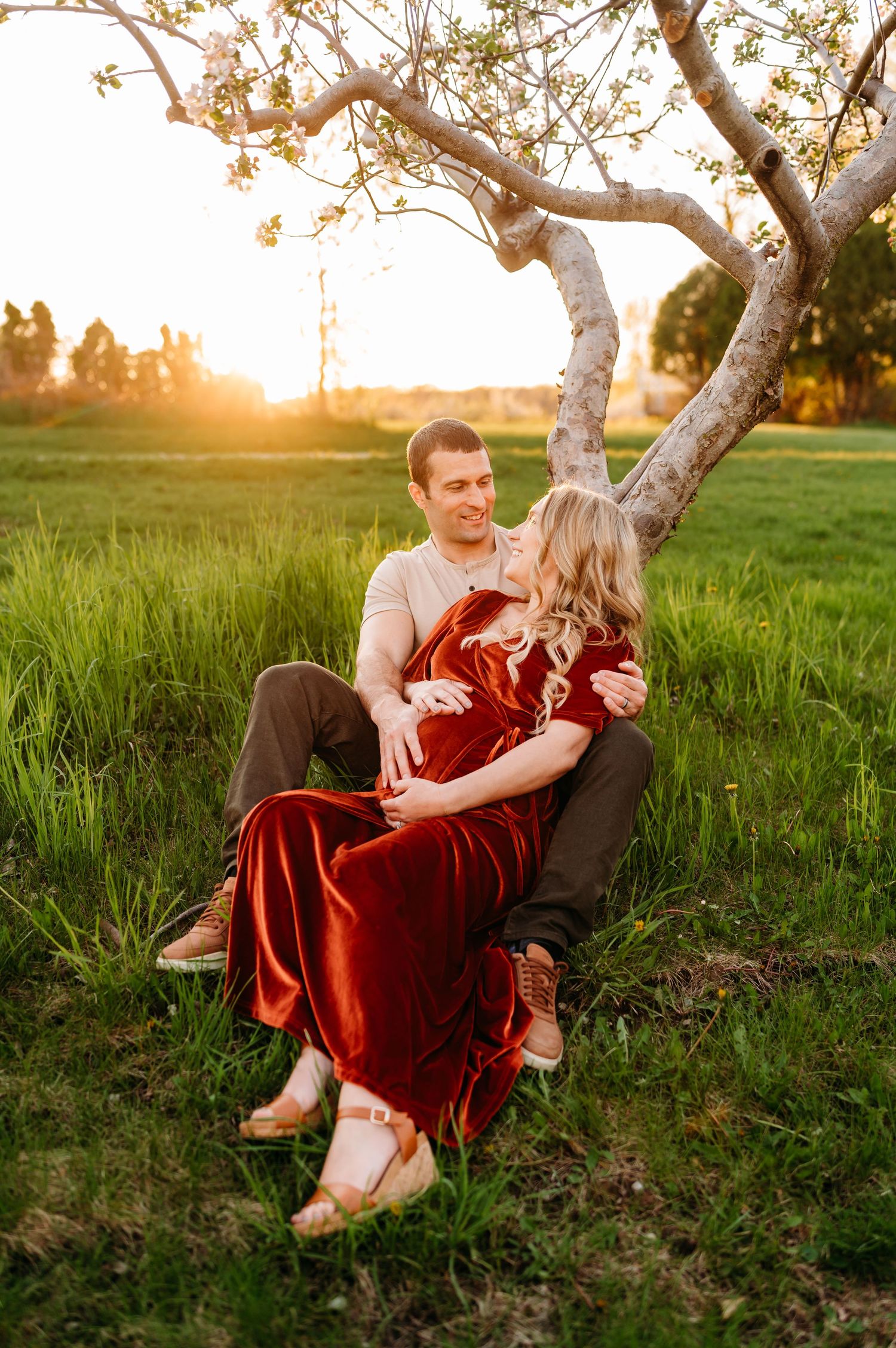 Couple sitting together under a curved tree at sunset in a grassy field with warm golden lighting.
