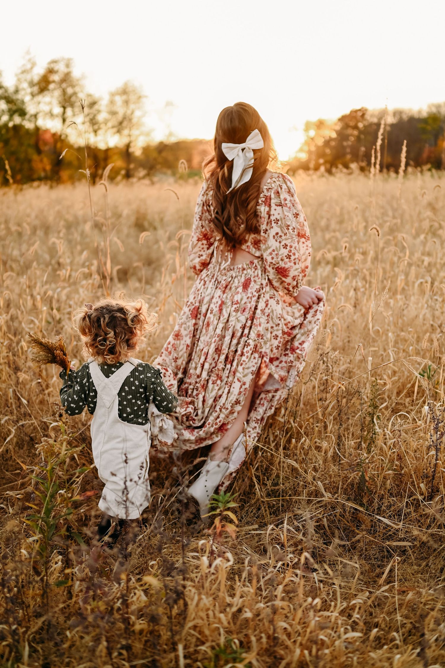 Mother and child in vintage-style clothing walk through a golden autumn field at sunset as their dresses sway in the breeze.