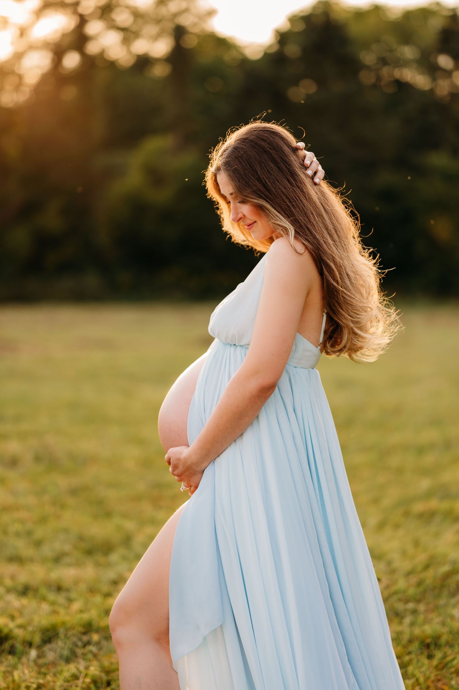 A pregnant woman in a flowing light blue dress poses in a sunlit field at golden hour.
