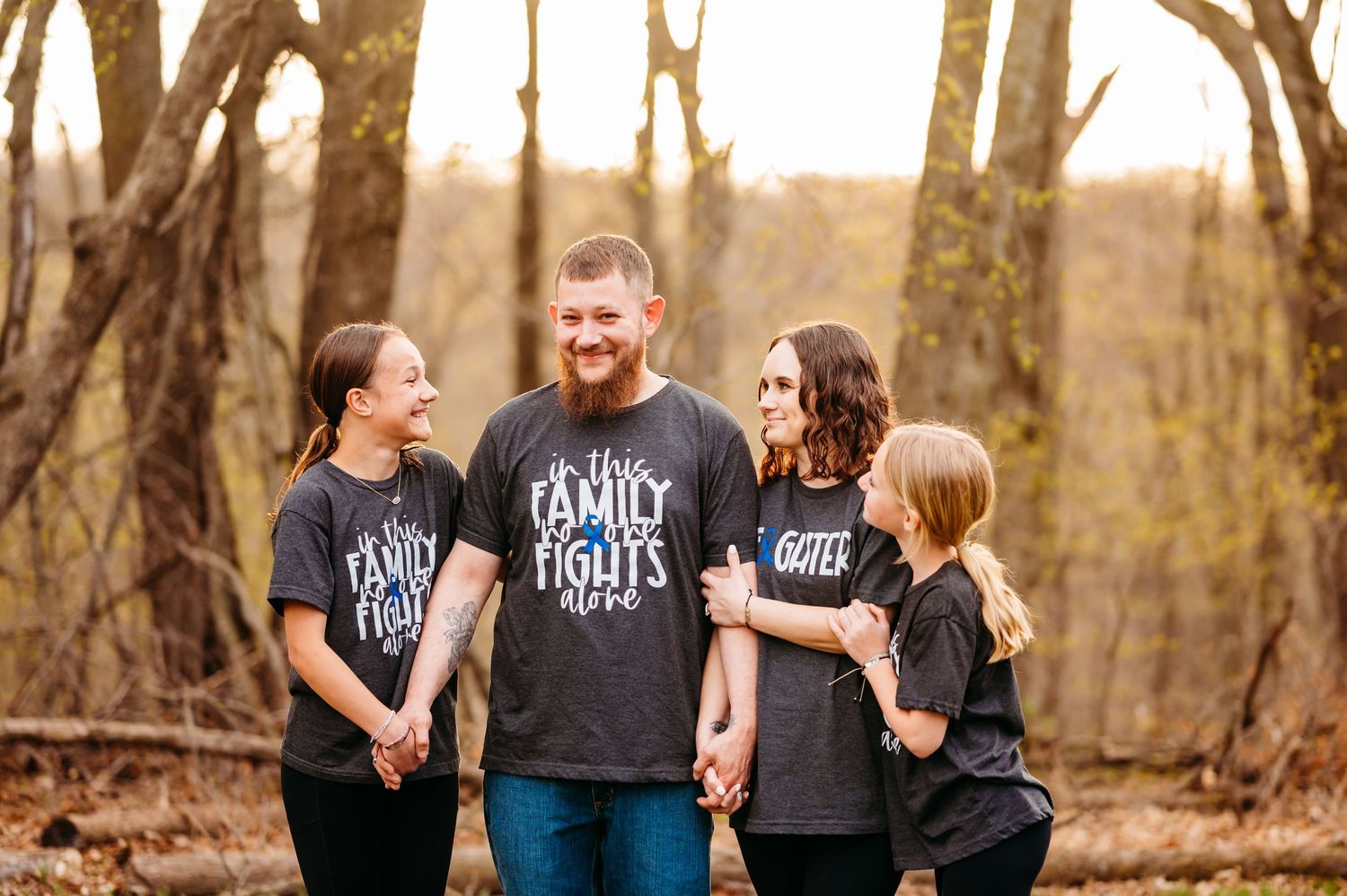 A family wearing matching black t-shirts laughs together while standing in a wooded area during golden hour.
