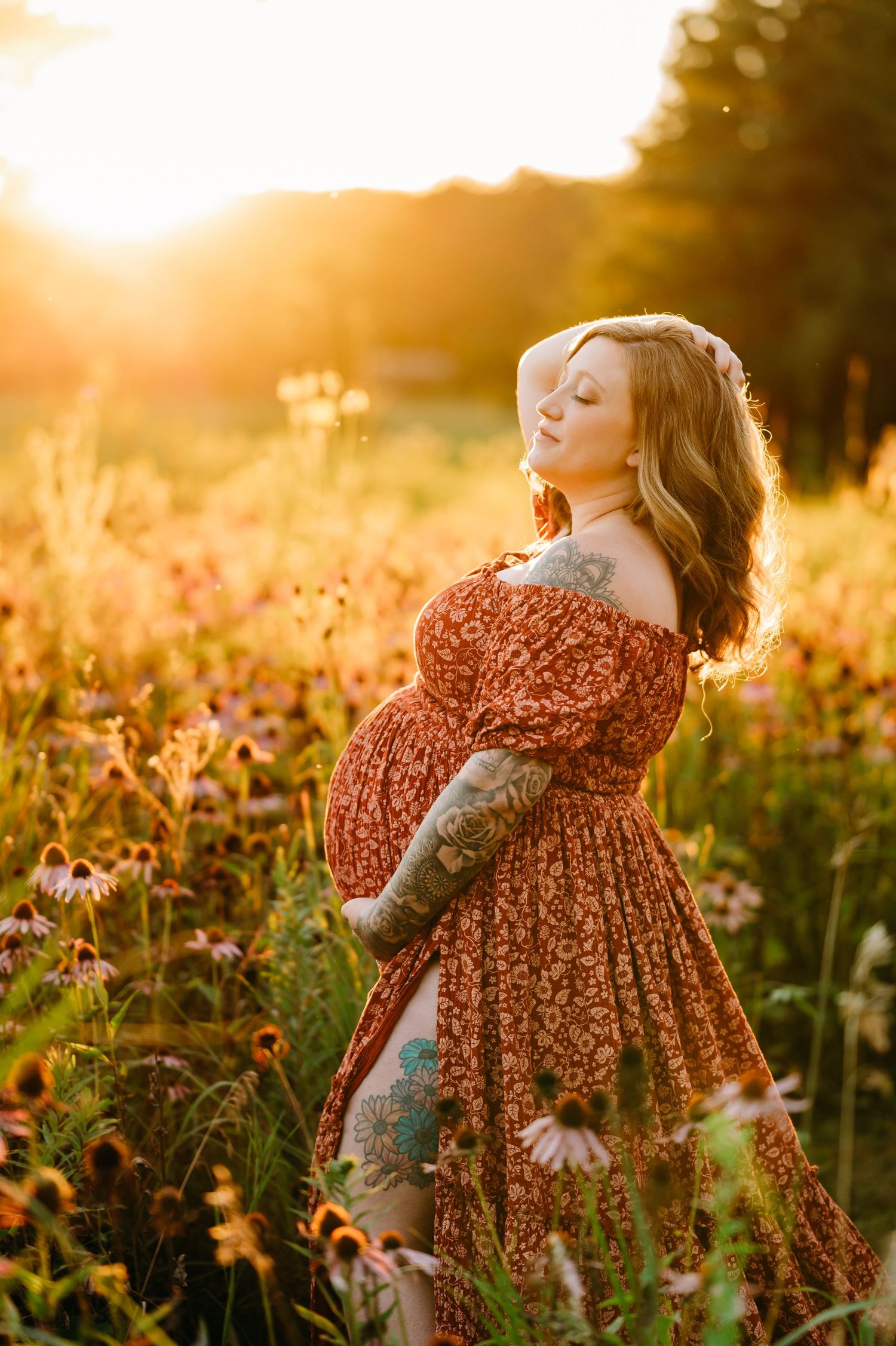 A pregnant woman in a floral dress stands in a golden sunlit field of wildflowers at sunset.