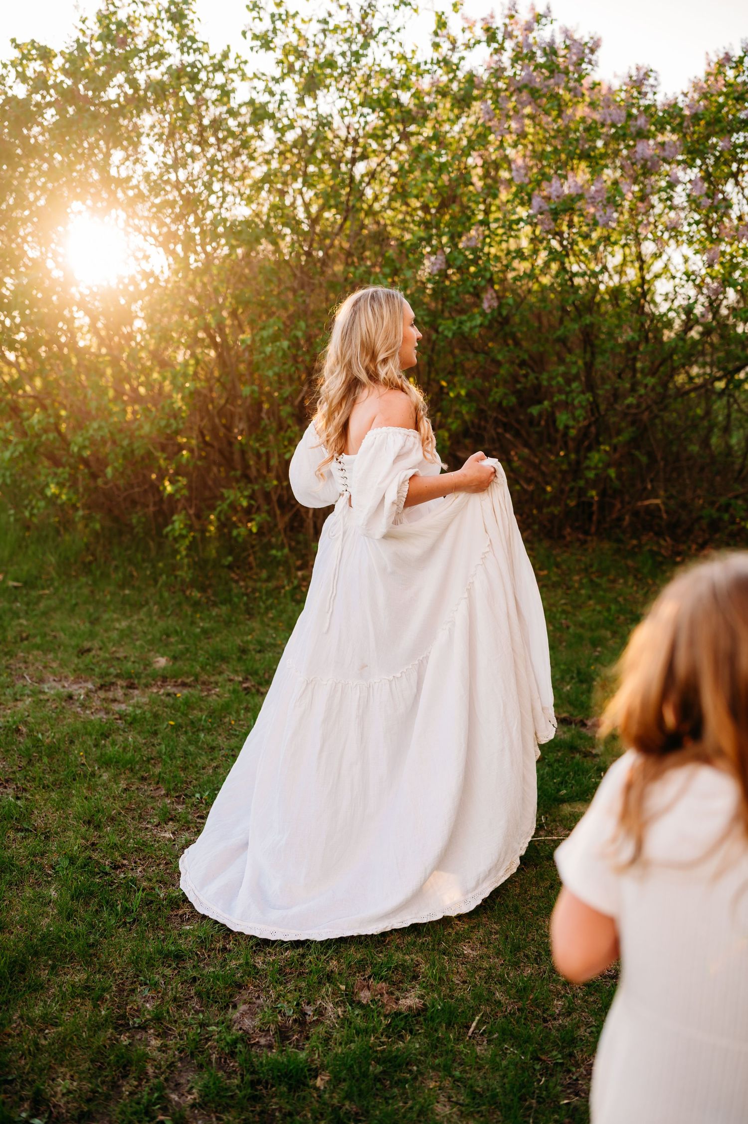 Person in flowing white off-shoulder dress twirls in garden at golden hour with sunlit trees and flowers in background.