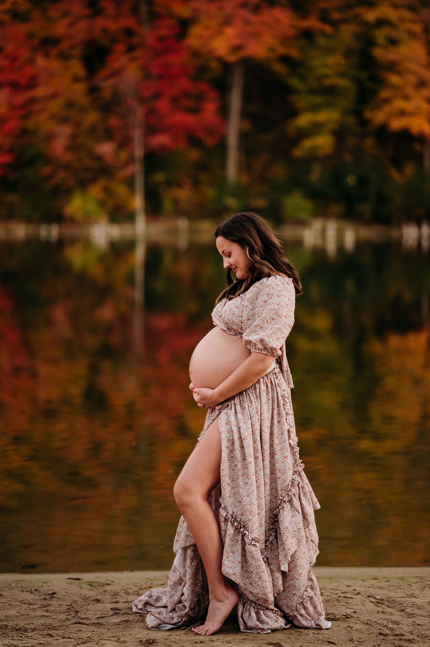 Pregnant woman in flowing lace dress poses near autumn lake with vibrant fall foliage reflecting on water.