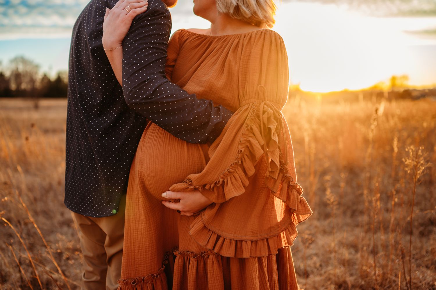 Two people wearing flowy dresses embrace in a golden sunlit field during sunset.