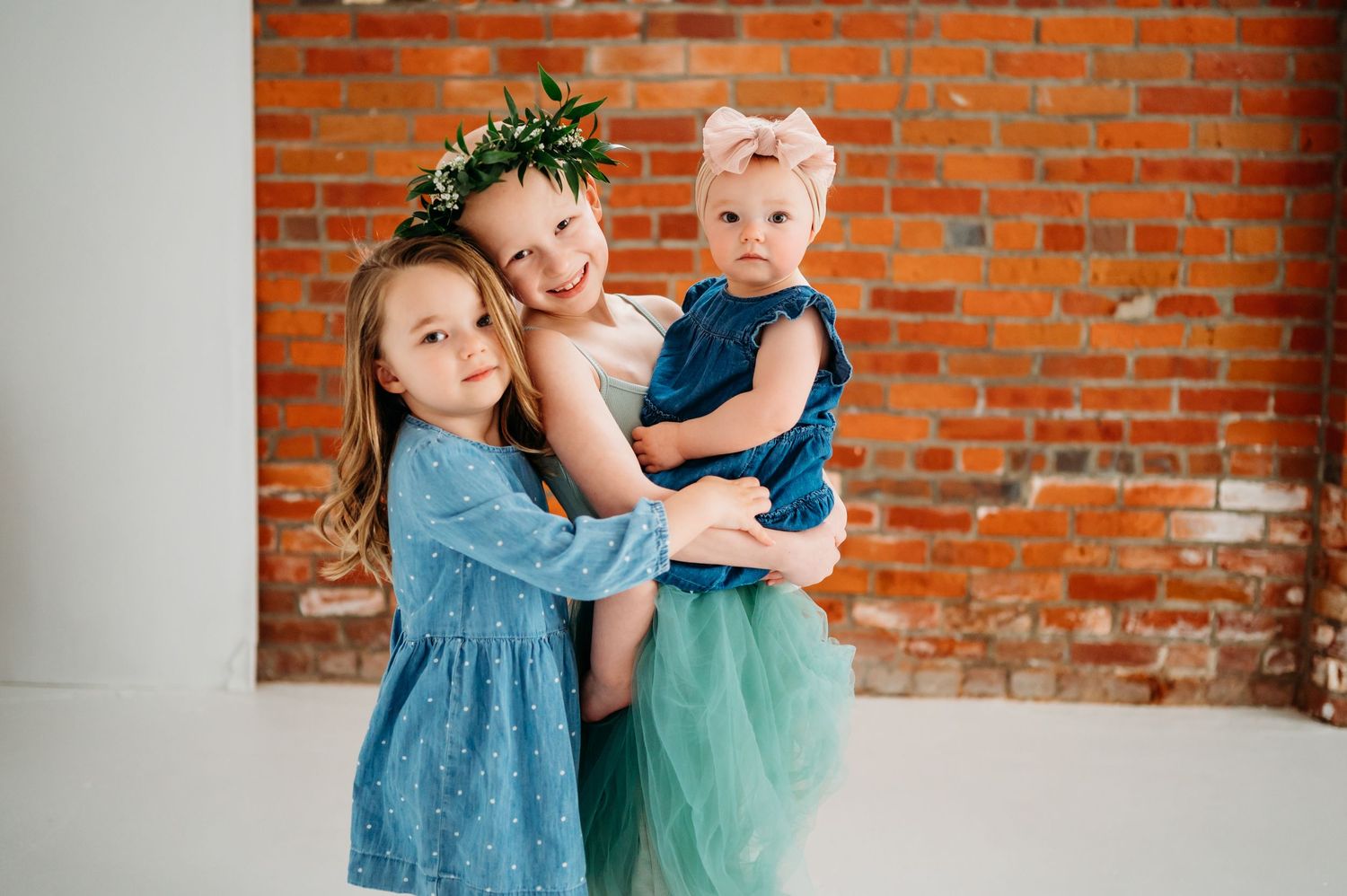 Two children in blue and mint green dresses embrace while standing against a rustic brick wall.