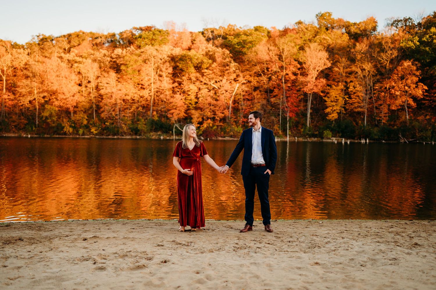 A couple stands together during a fall photoshoot near a lake with vibrant orange foliage in the background.