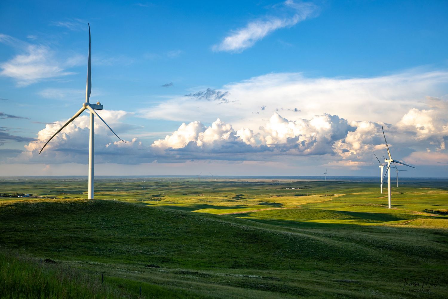 Wind turbines stand tall over rolling green hills beneath a dramatic cloudy sky in a vast open prairie landscape.