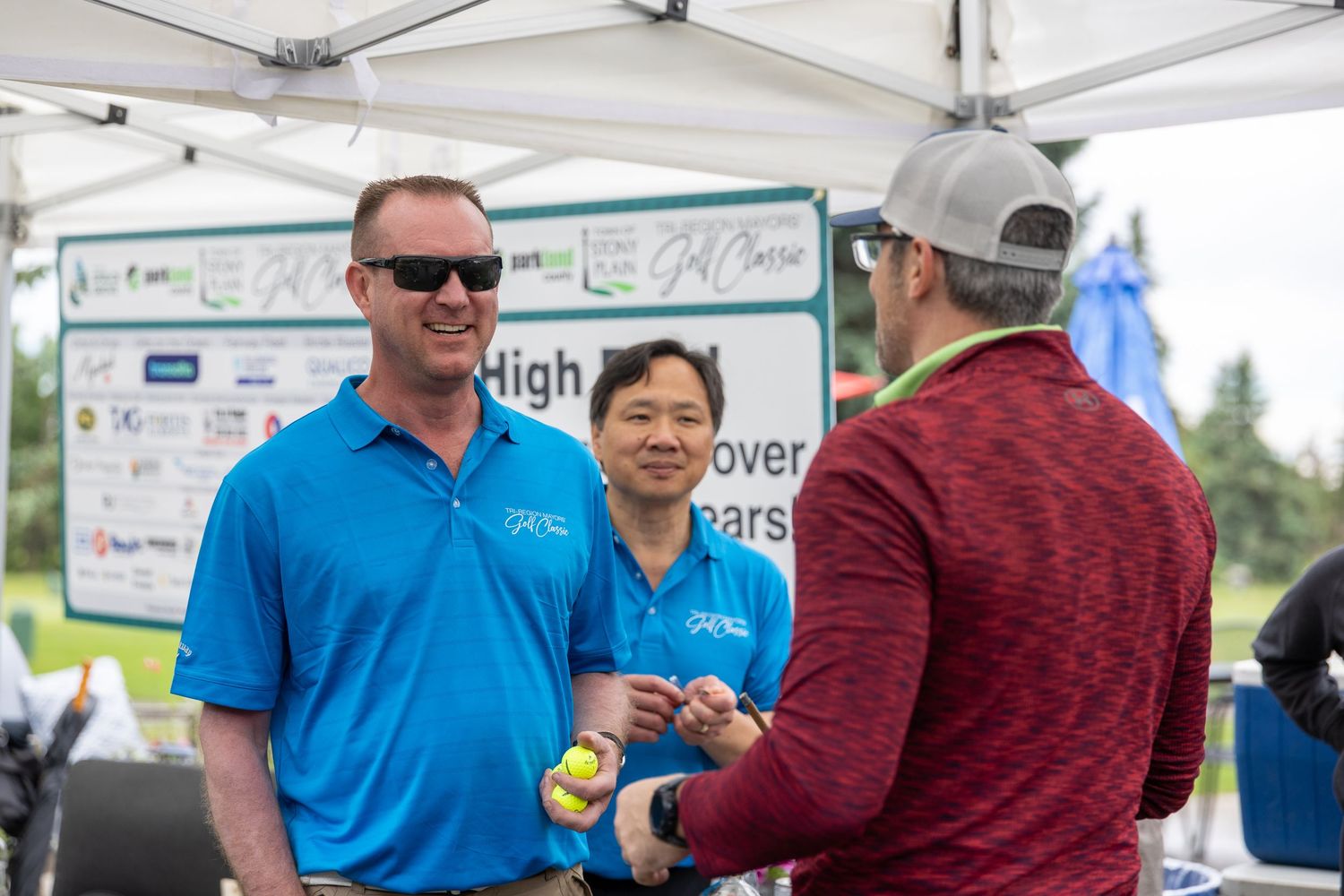 Three people networking at a golf course event tent, two wearing blue polo shirts, exchanging items outdoors.