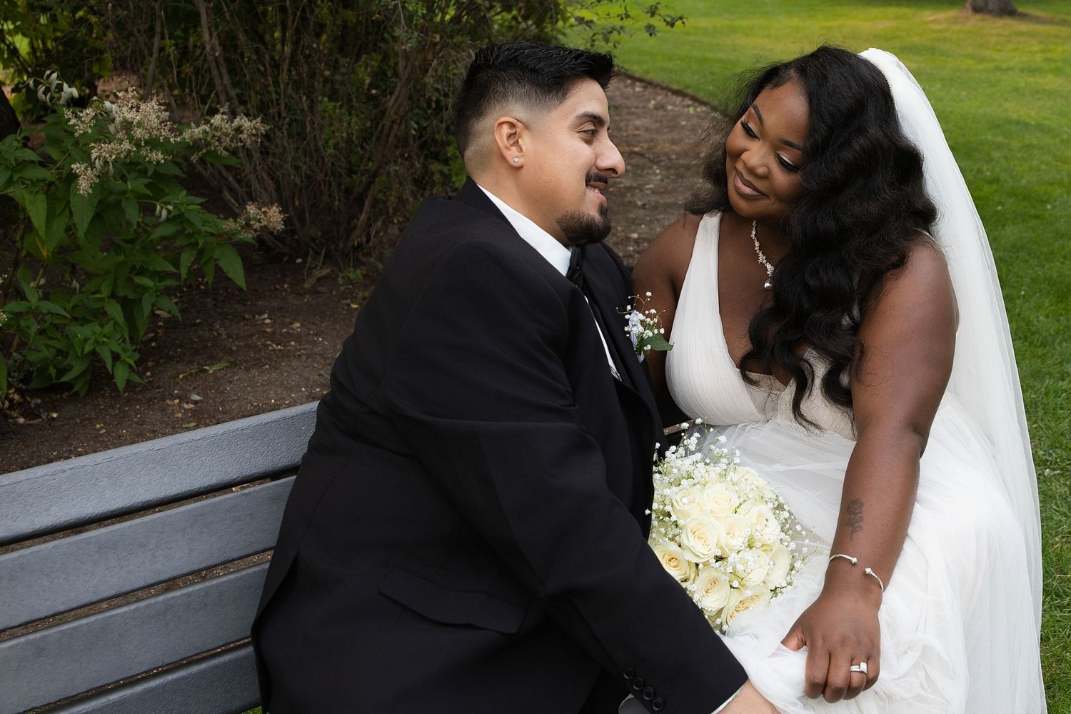 Bride and groom sitting on a bench, smiling at each other, bride holding white bouquet in outdoor wedding setting.