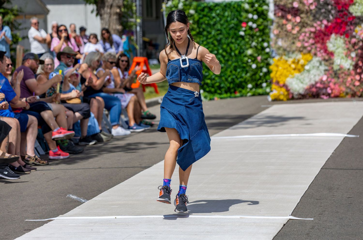Young woman in a blue dress walks confidently down an outdoor fashion runway with a crowd of spectators.
