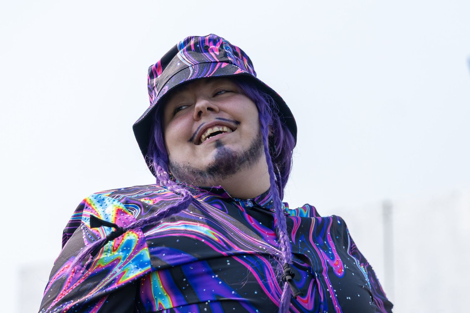 Person wearing a vibrant purple and multicolor holographic bucket hat and matching outfit, smiling outdoors.
