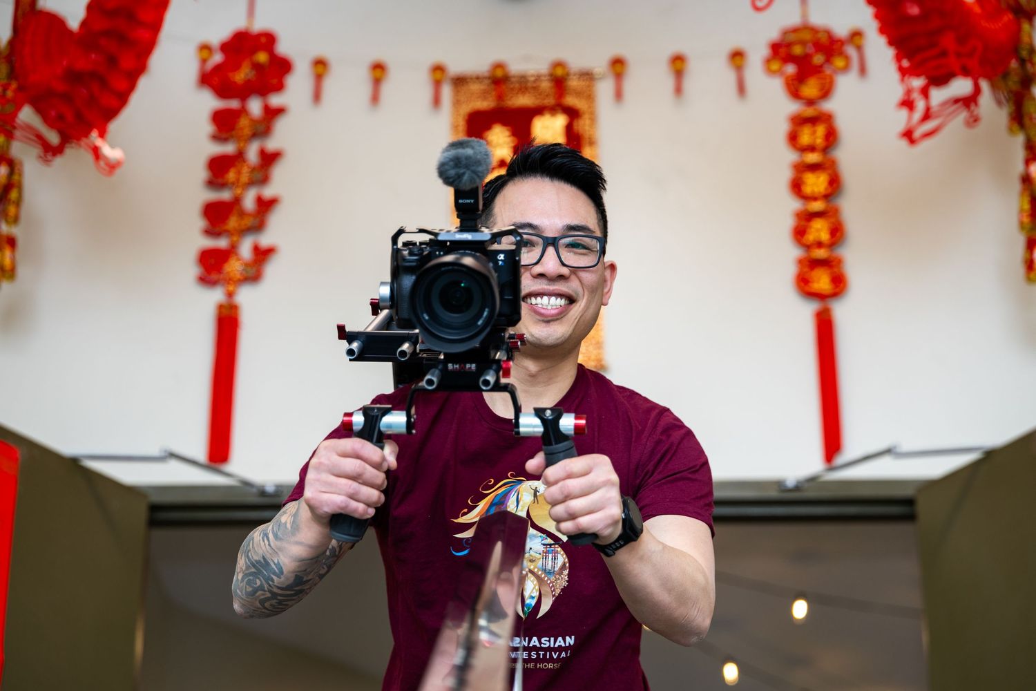 Smiling Asian man holding a professional camera on a gimbal stabilizer, with Chinese New Year decorations in the background.