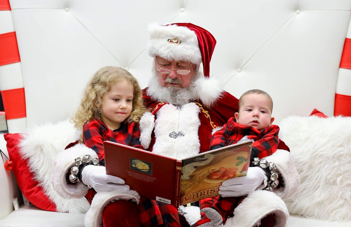 Santa Claus reading a Christmas book with two young children in festive red plaid outfits on a white chair.
