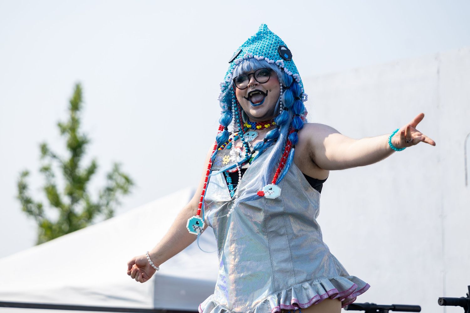 Person in colorful blue wig and silver costume dancing on stage at outdoor festival.