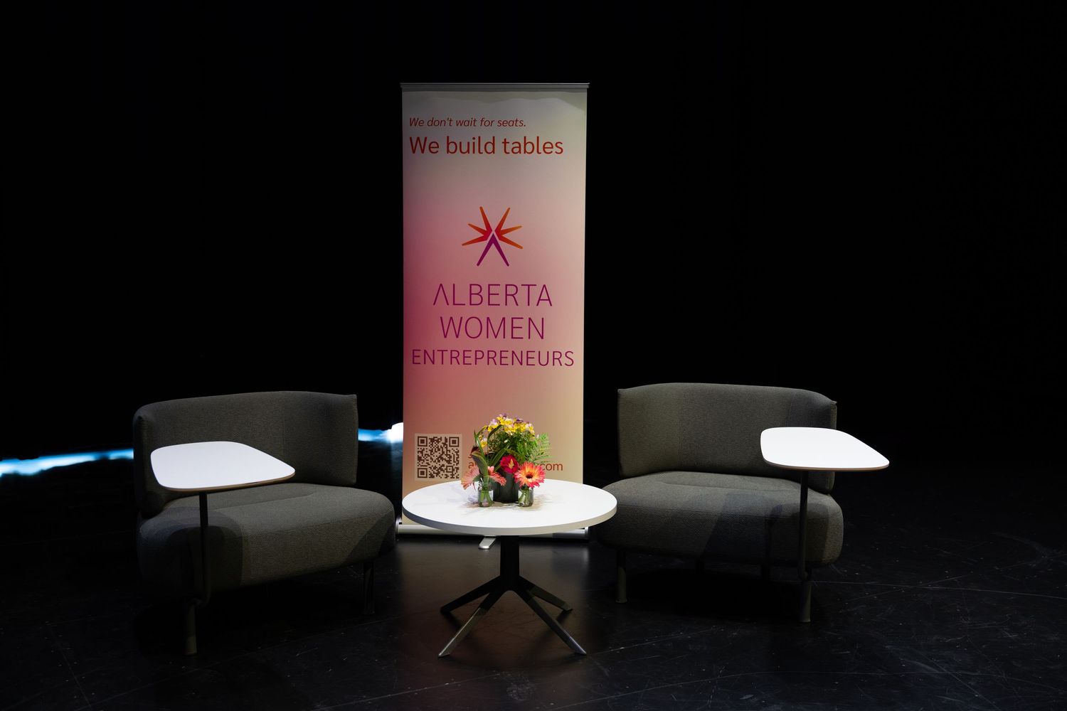 Two chairs and a small table set up in front of an Alberta Women Entrepreneurs banner on a dark stage.