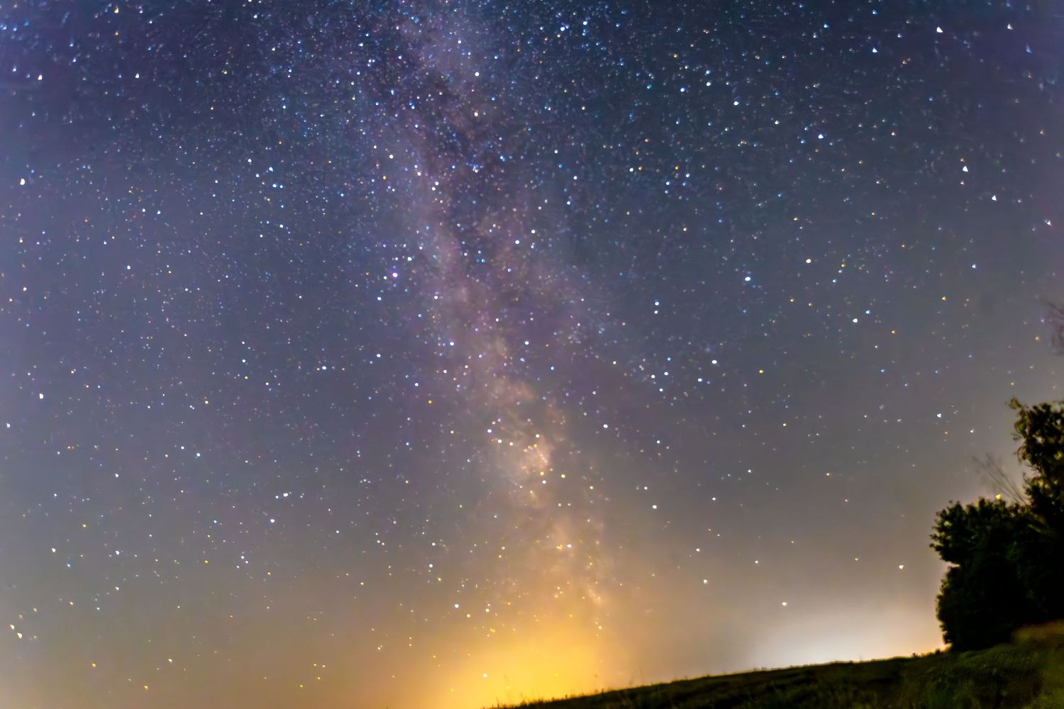 Stunning Milky Way galaxy stretches across a star-filled night sky above a dark silhouetted hillside landscape.