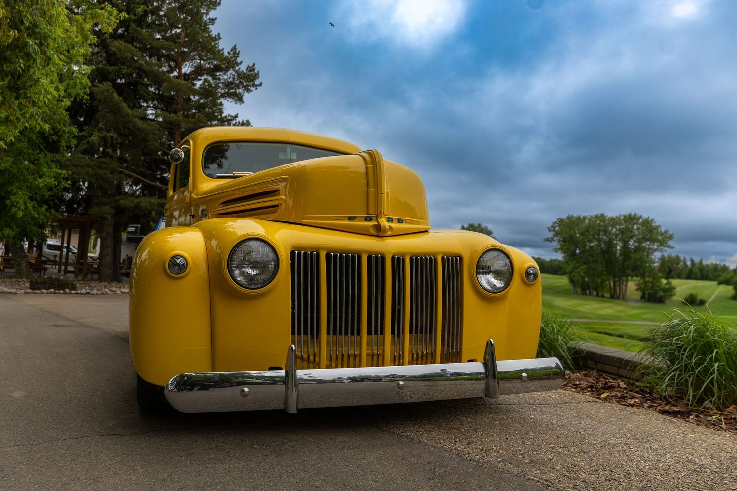 Vintage yellow Ford truck parked on a path with lush green trees and cloudy sky in the background.