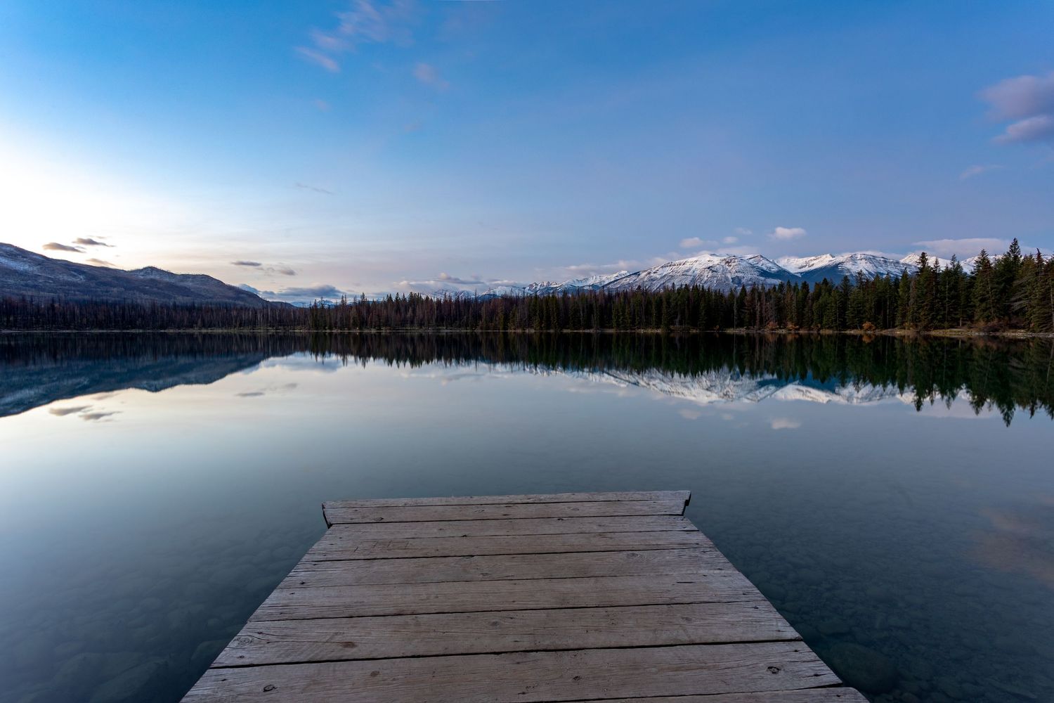 Wooden dock extending into a calm reflective mountain lake with snow-capped peaks and evergreen trees at dusk.