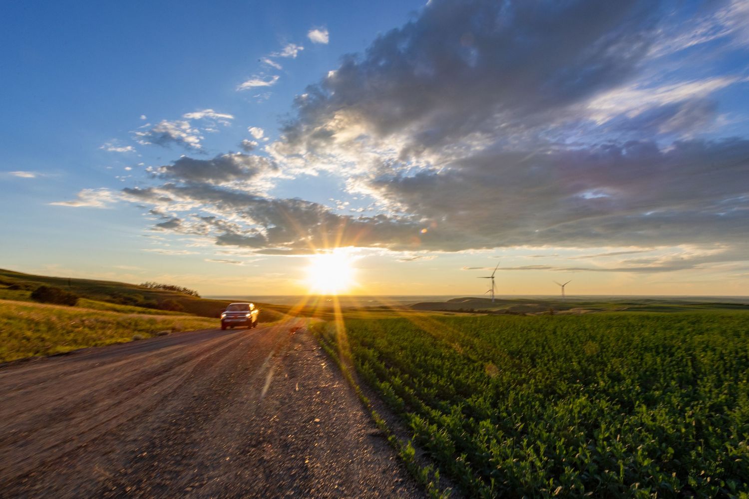 Car driving on a dirt road through green fields at sunset with wind turbines and dramatic clouds in the sky.
