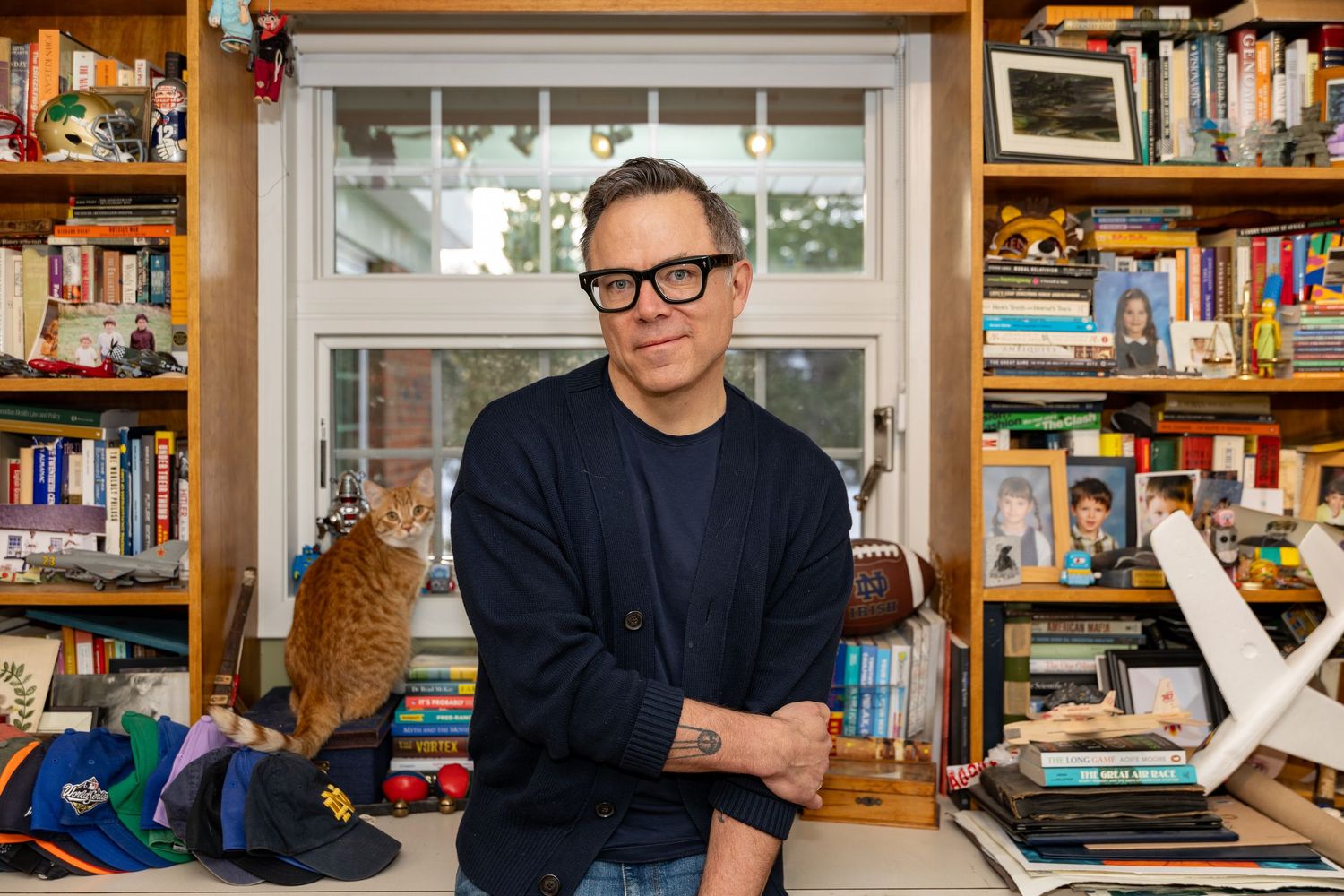 Man with glasses sits smiling before bookshelves with an orange cat, in a cozy home library setting.