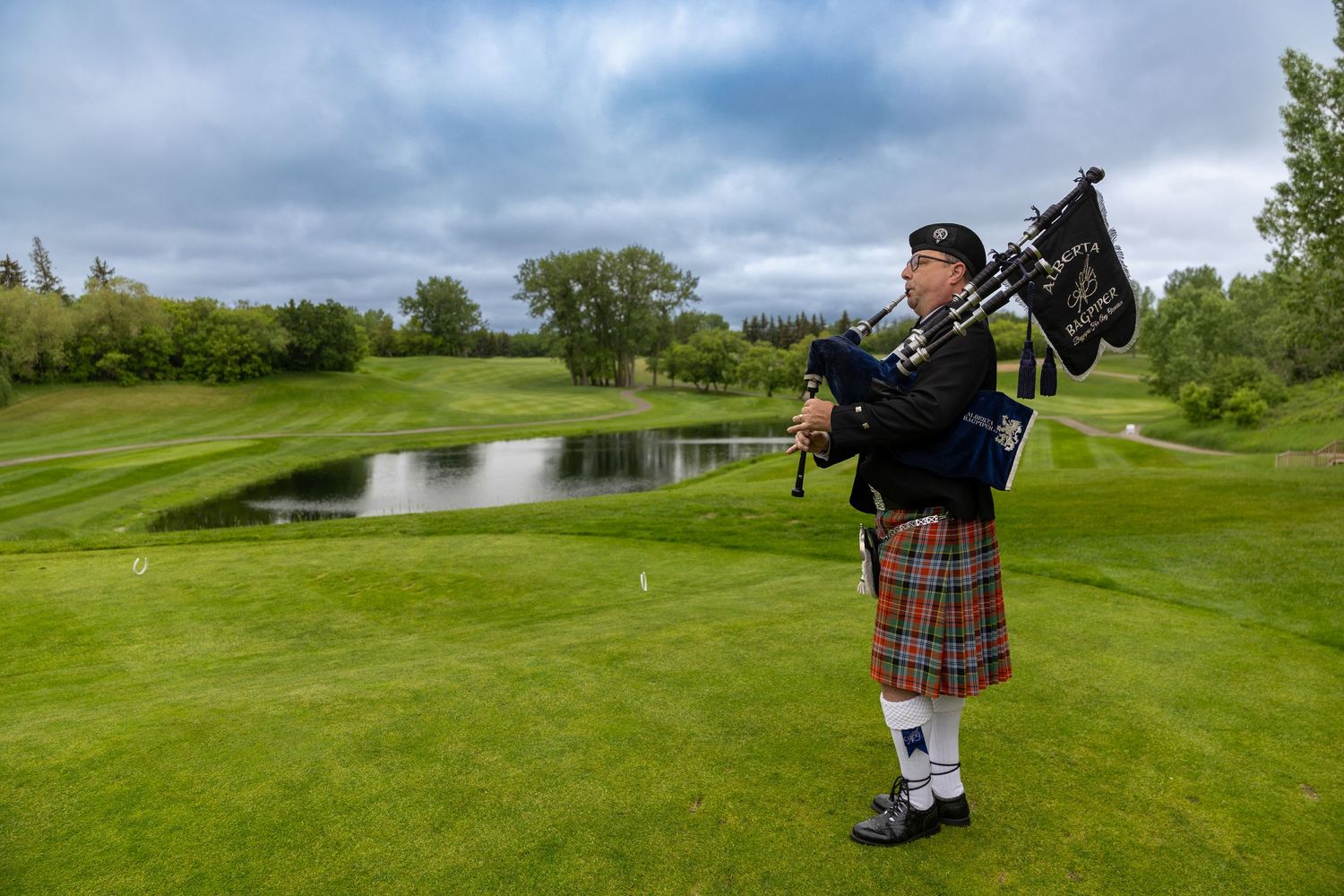 Bagpiper in traditional Scottish kilt playing on a lush green golf course with a pond and trees in the background.