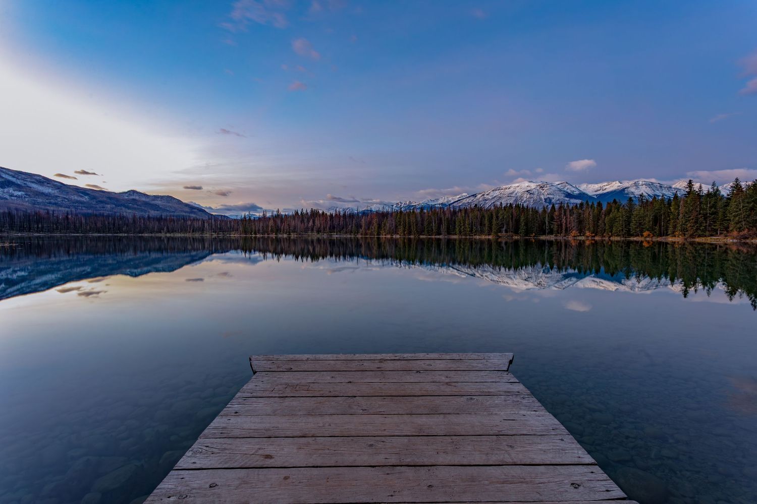 Wooden dock leads over a mirror-like mountain lake reflecting snow-capped peaks and evergreen forest at dusk.