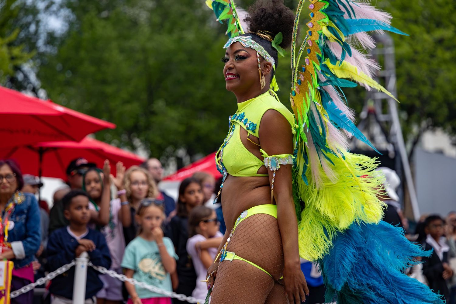 A woman in vibrant neon yellow carnival costume with colorful feathers performs at a lively outdoor festival parade.