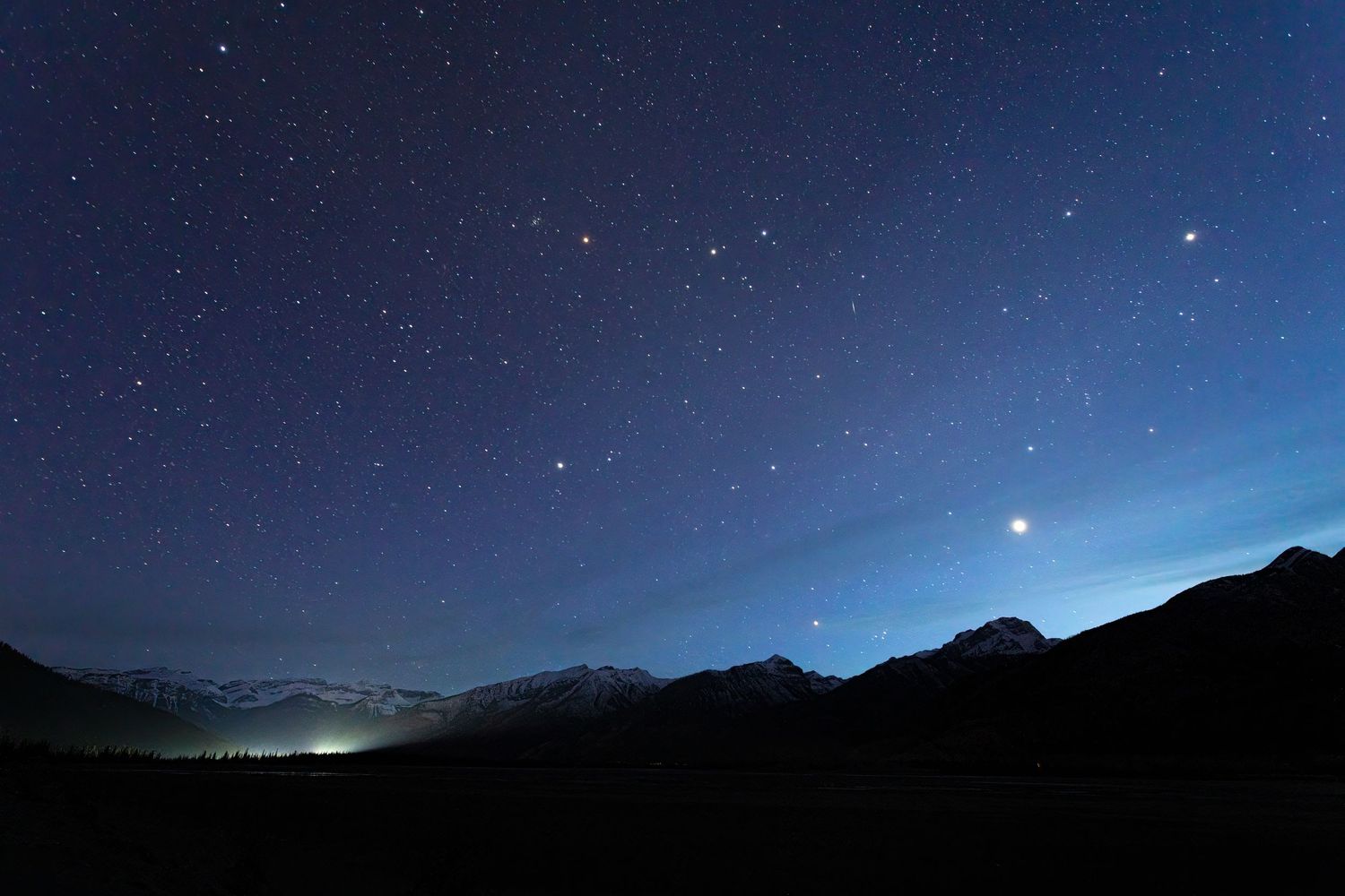 Starry night sky over silhouetted mountain range with bright stars and glowing horizon light.