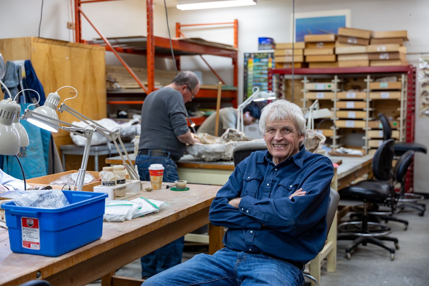 Man in denim shirt sits arms crossed smiling in a busy workshop with shelves, workbenches, and a colleague working in background.