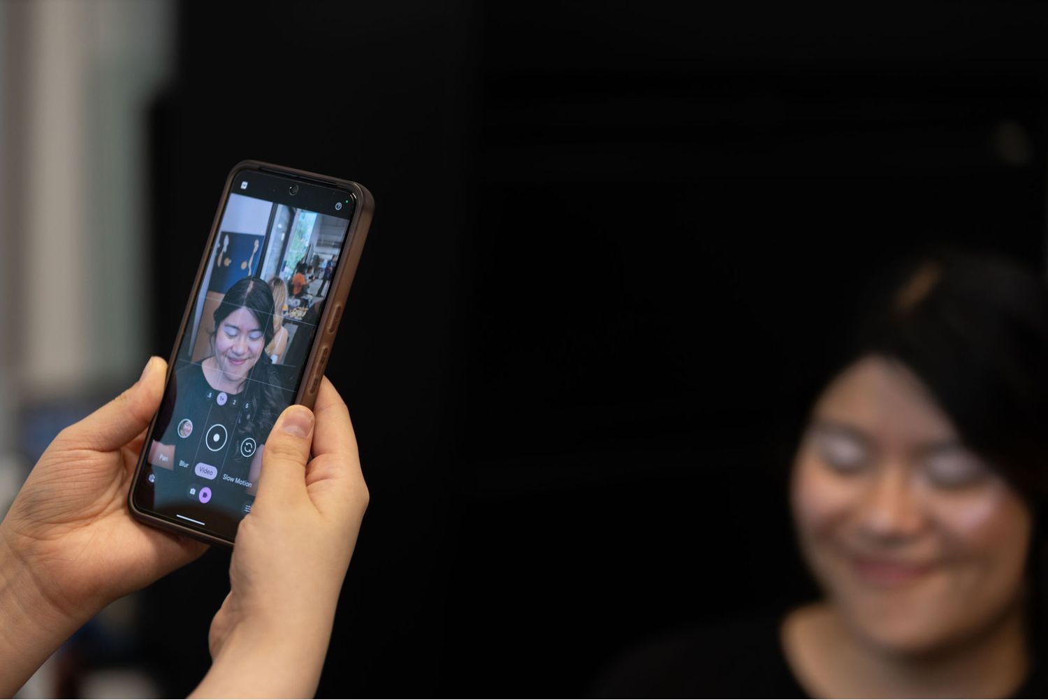 Person photographing a smiling woman with a smartphone, capturing her portrait on the phone screen.