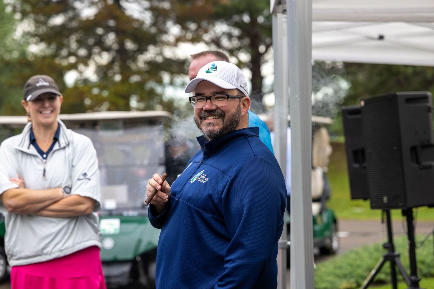 Man in blue jacket smiling at golf event, holding microphone, with woman in pink pants and golf cart in background.