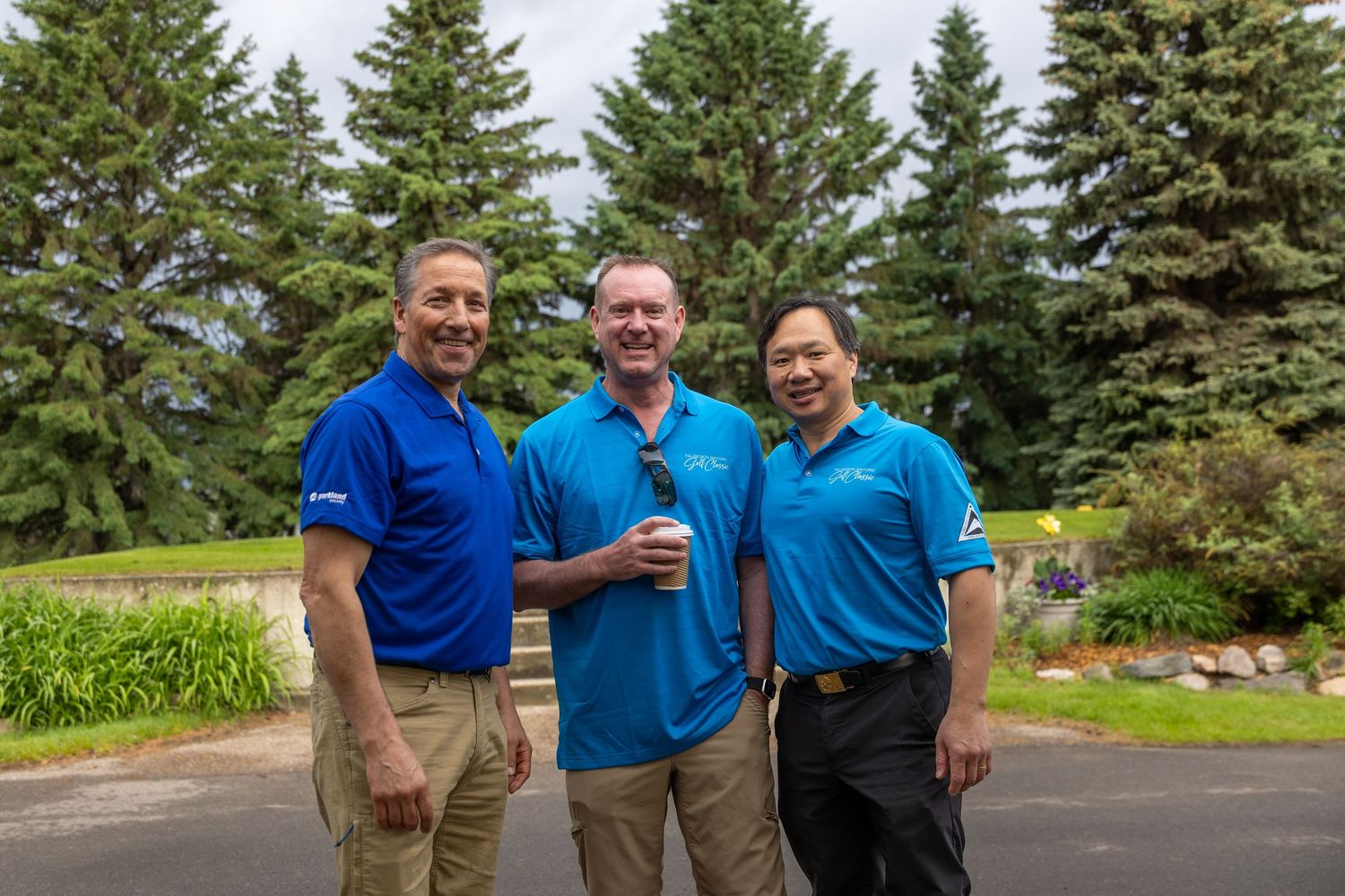Three men in blue polo shirts smiling outdoors near evergreen trees at a golf event.