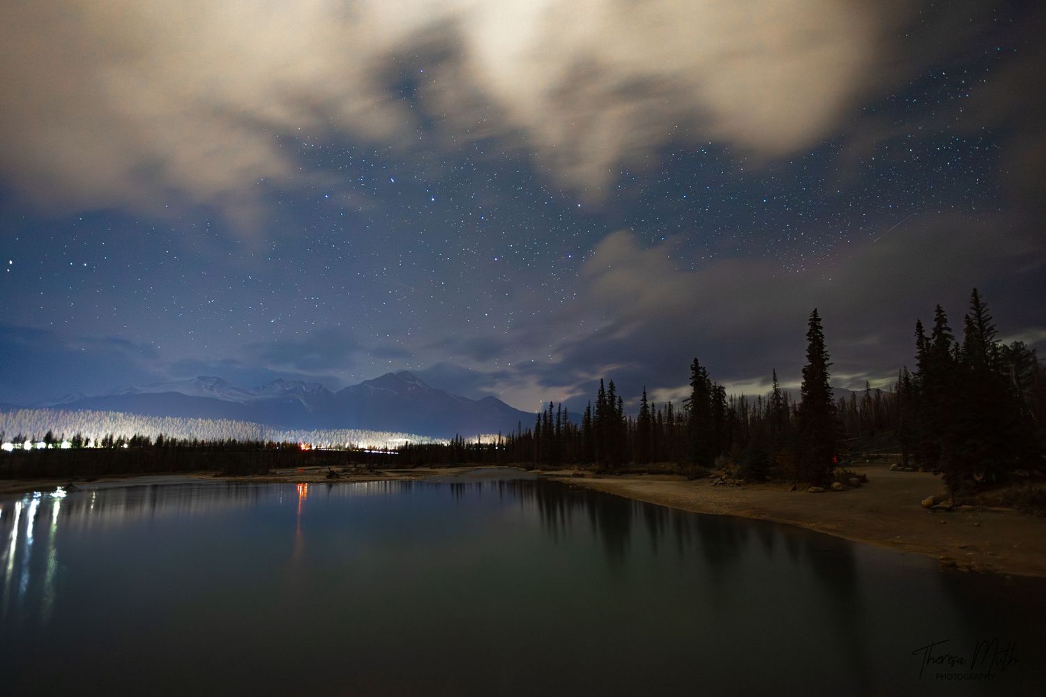 Starry night sky over a calm river with distant town lights reflecting on the water among pine trees.