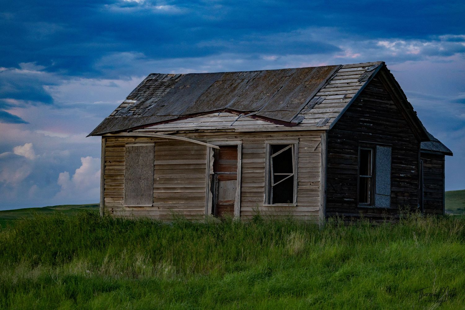 Abandoned weathered wooden farmhouse on green prairie under dramatic stormy blue sky.