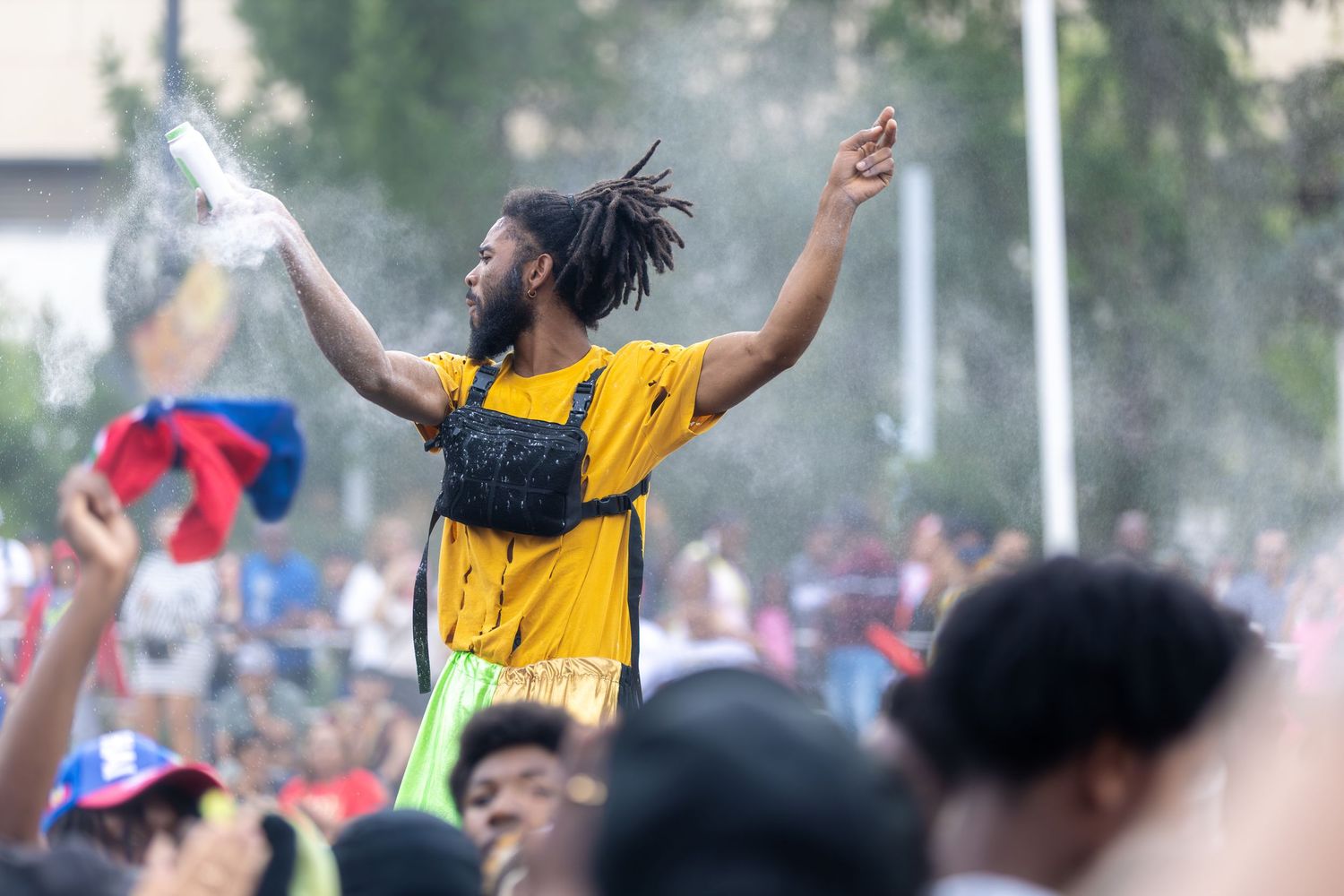 Man with dreadlocks in yellow shirt raises arms in crowd at outdoor summer music festival.