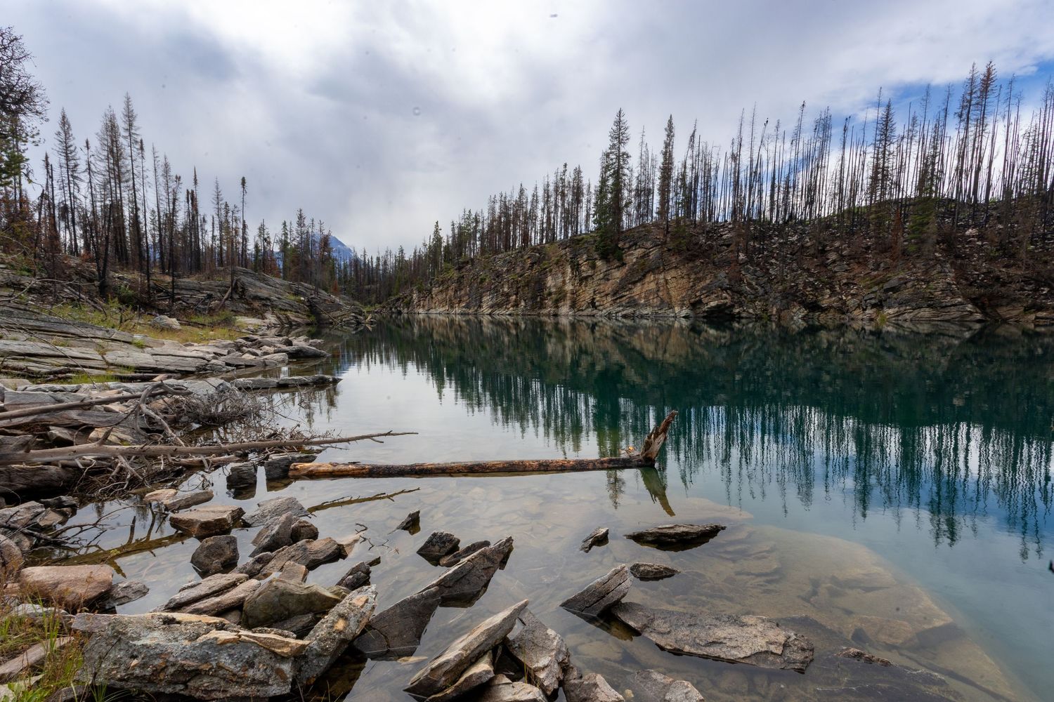 Serene turquoise lake reflecting burnt forest and rocky shoreline under dramatic cloudy sky.