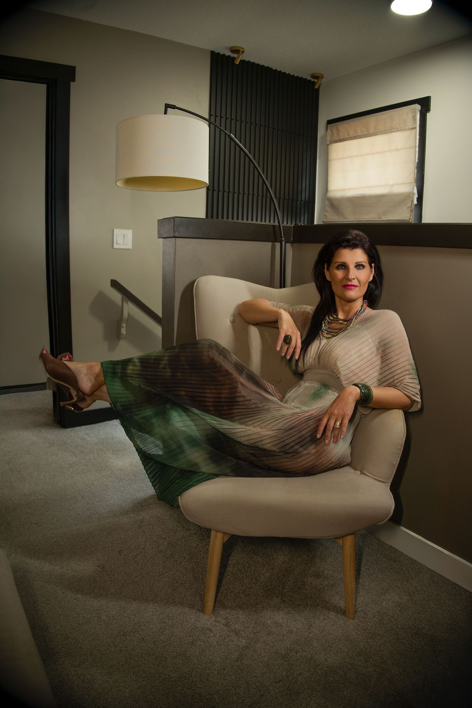 Woman in green skirt and beige top relaxing in modern chair with floor lamp in stylish room.