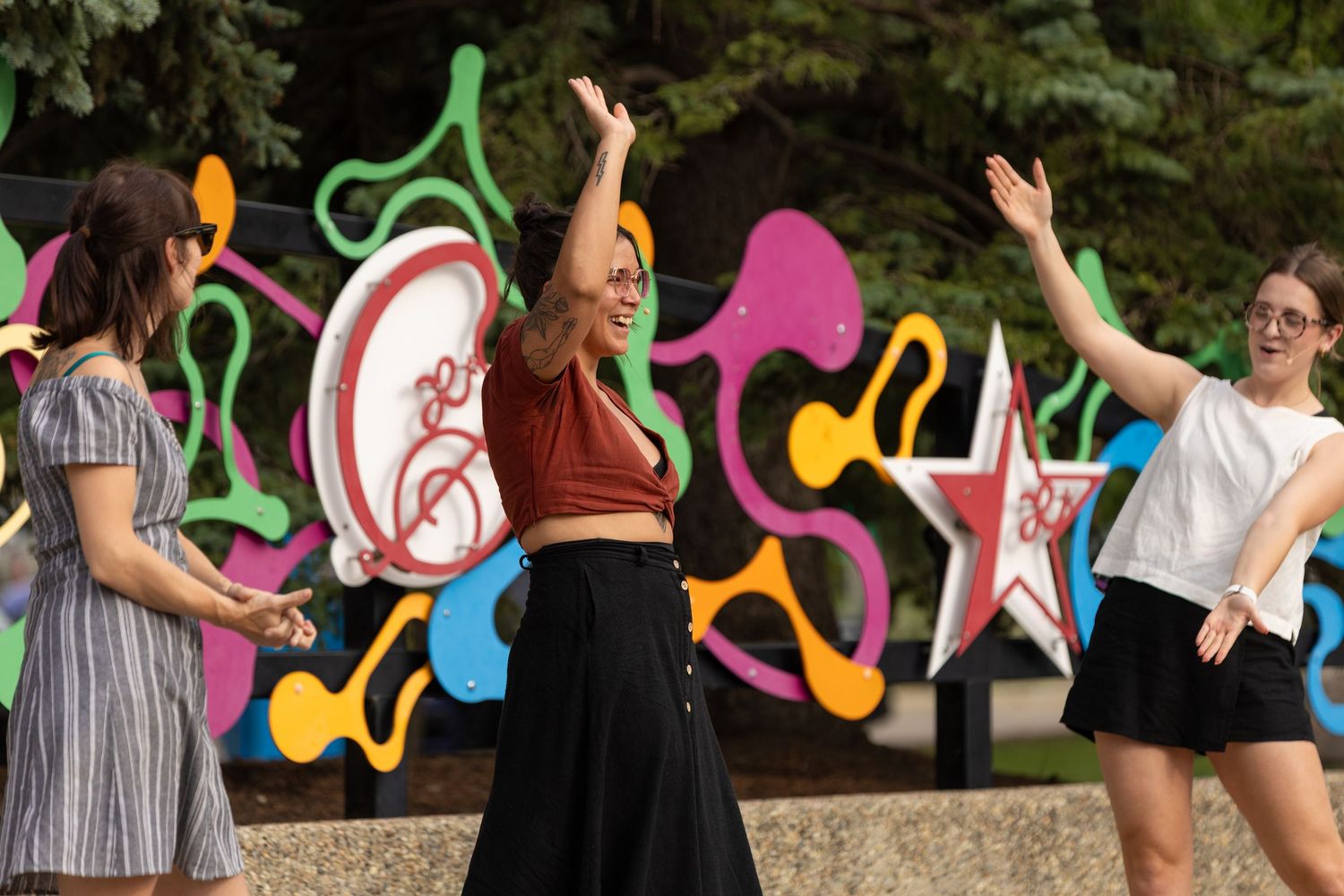 Three women dance joyfully outdoors in front of a vibrant colorful art installation on a sunny day.