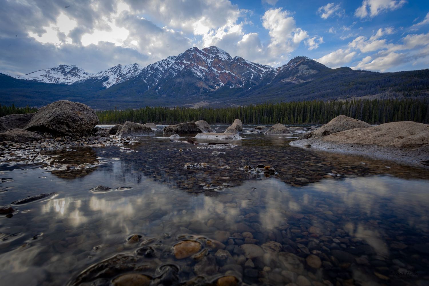 Rocky mountain stream reflects snow-capped peaks and dramatic clouds in a pristine wilderness landscape.