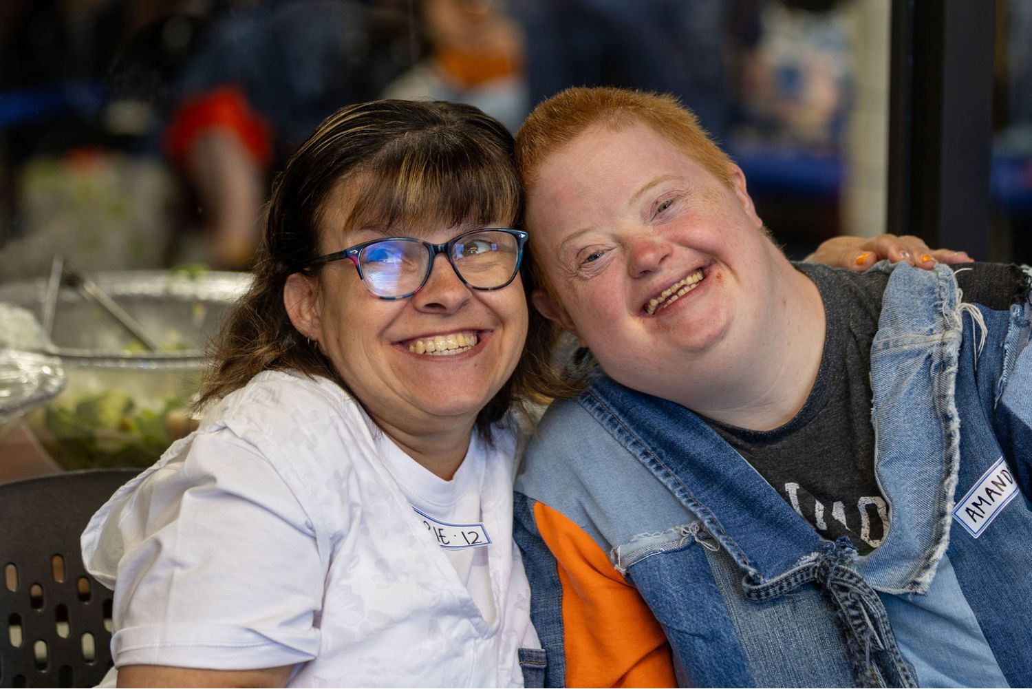 Two smiling women pose together at an event, one wearing glasses and a white shirt, the other in a denim jacket.
