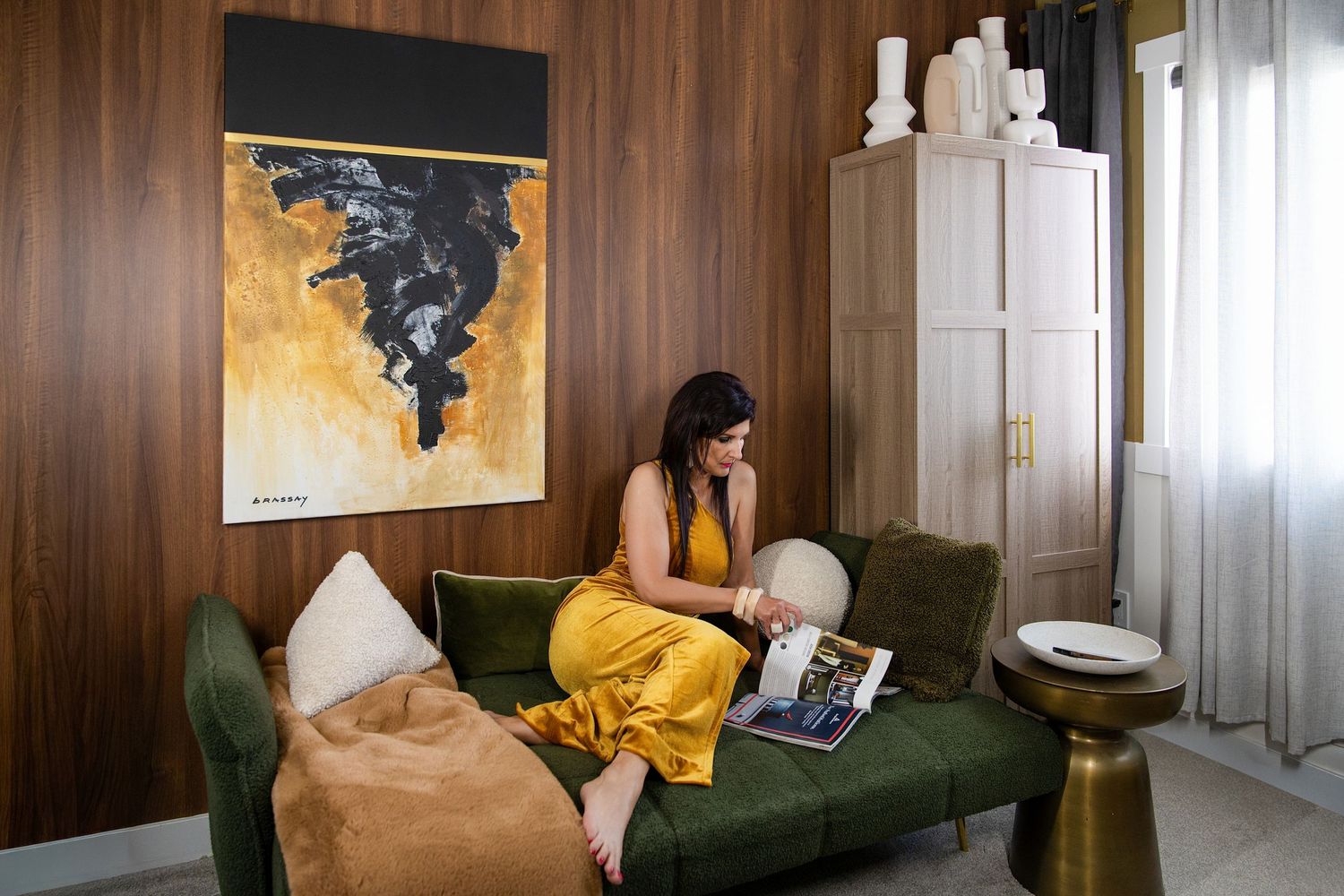 Woman in yellow dress reading on green velvet sofa beside abstract gold artwork in stylish wood-paneled room.