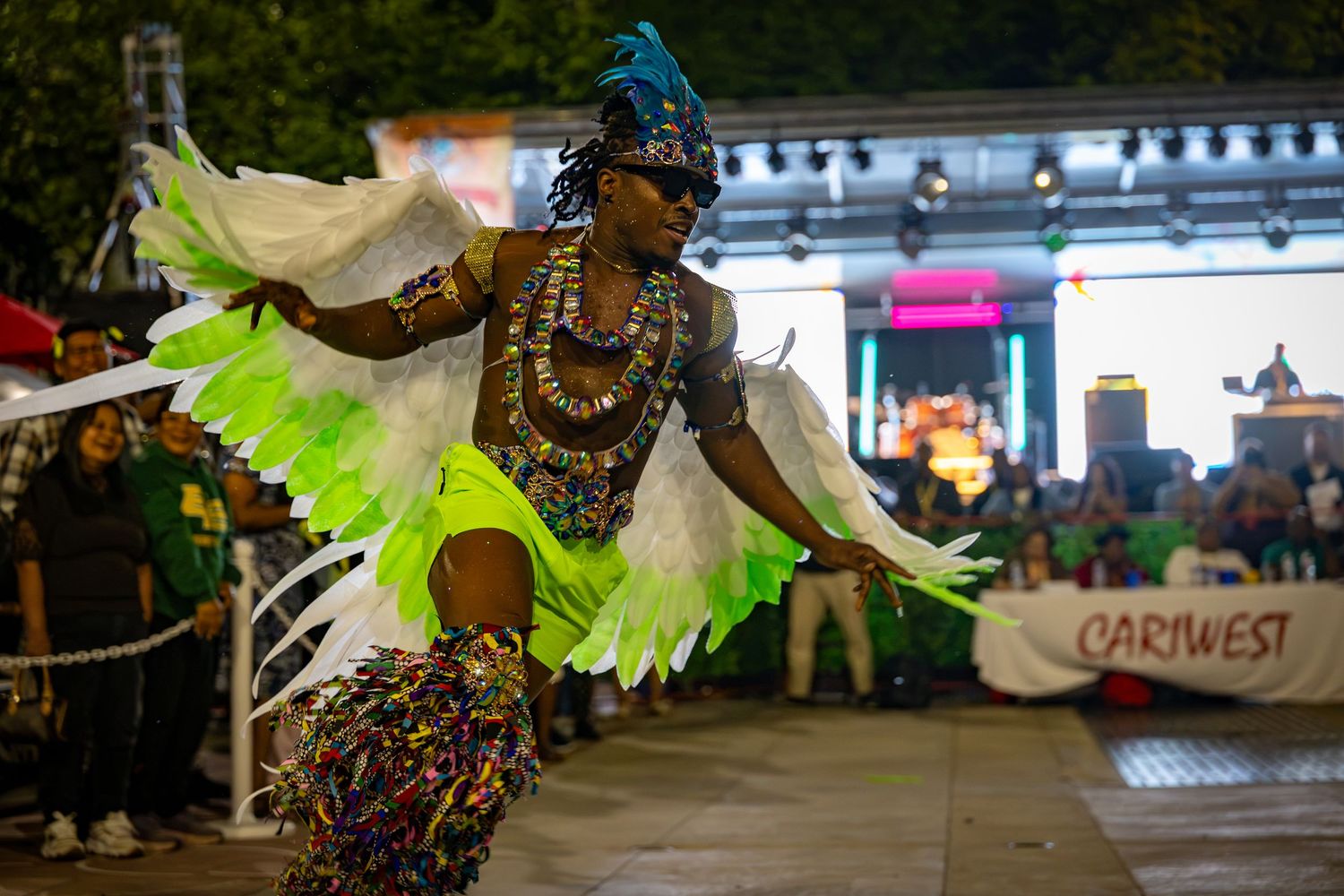 Dancer in vibrant green feathered costume performs at Cariwest Caribbean festival parade at night.