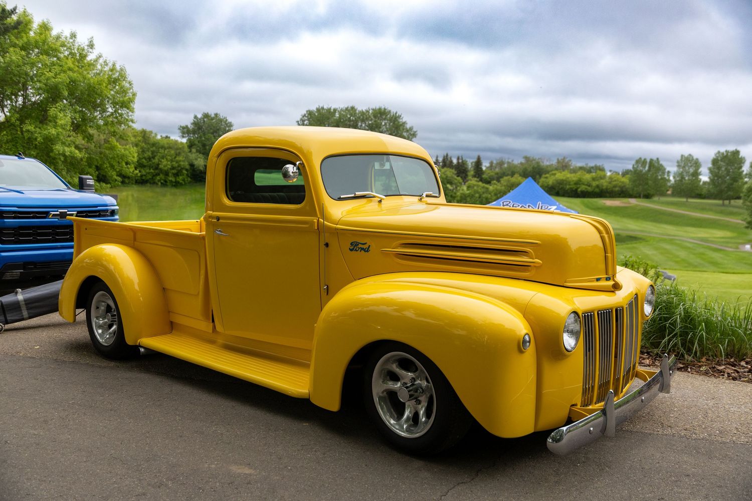Restored vintage yellow Ford pickup truck at a classic car show, parked on pavement with green trees in background.