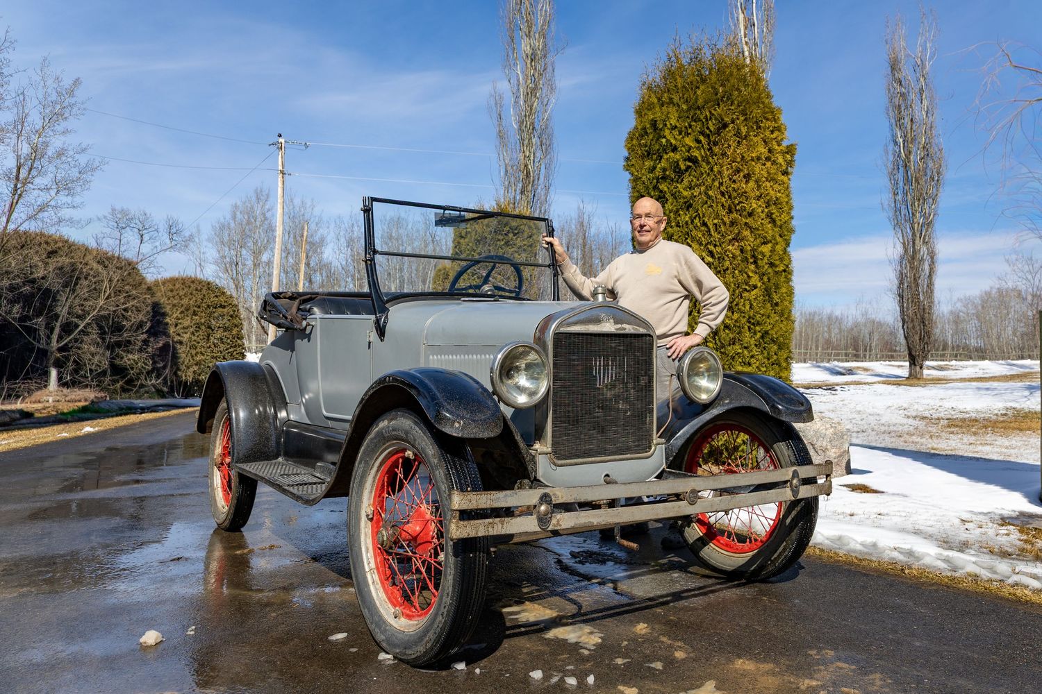 Man posing proudly beside a restored vintage gray Model T Ford with red wheels on a sunny winter day.