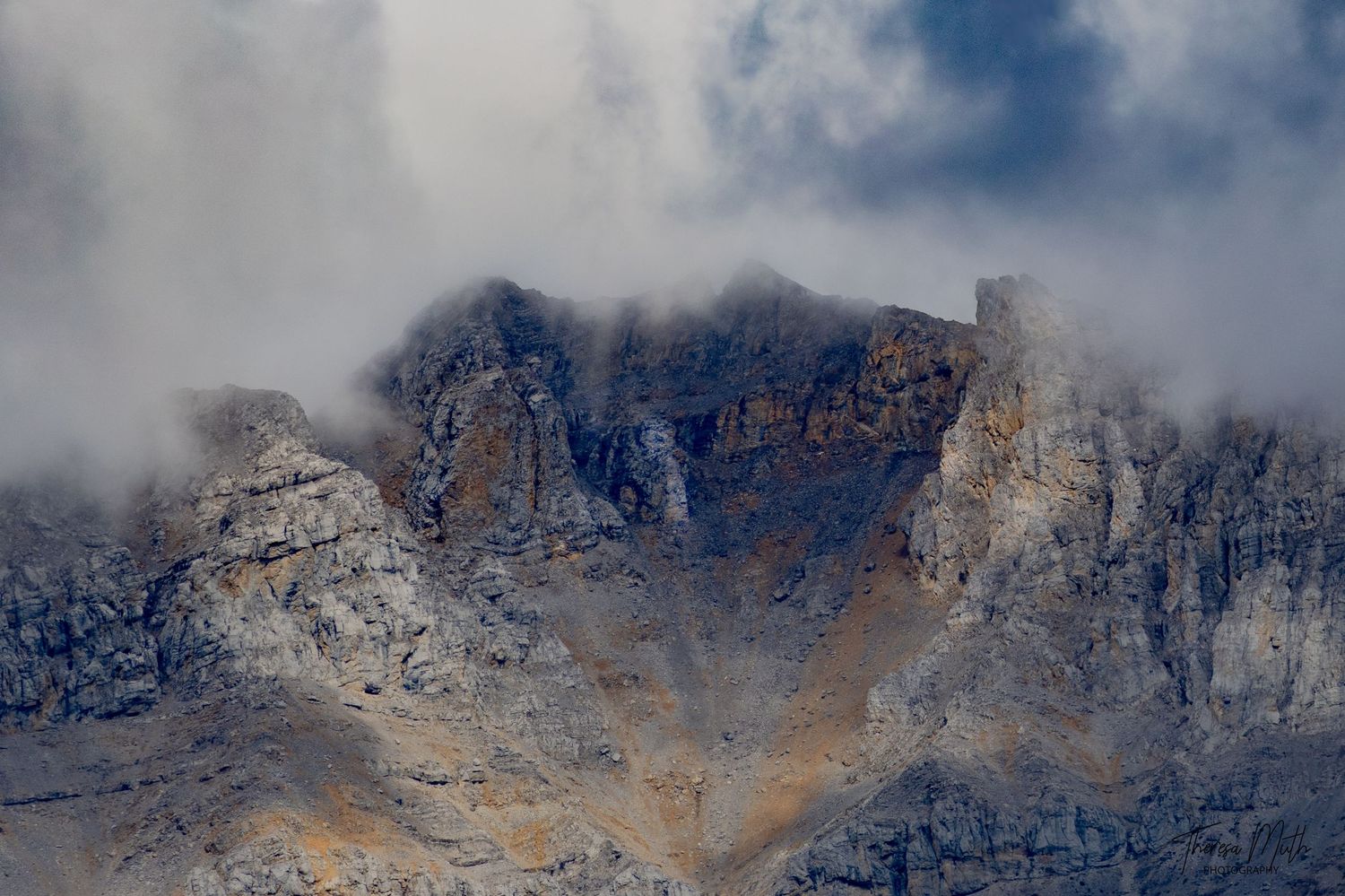 Dramatic rocky mountain peaks partially shrouded in thick clouds and mist against a grey sky.