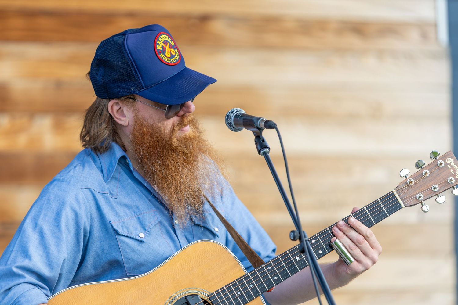 Bearded male guitarist performs at outdoor venue wearing blue cap and denim shirt.