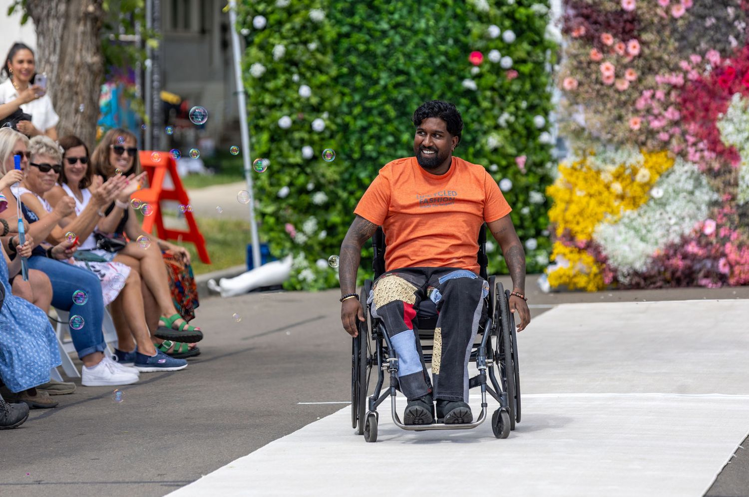 Man in an orange shirt uses a wheelchair on a fashion runway, smiling at the applauding audience.